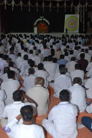 Large seated audience of men in white shirts and white dhotis fills a hall, facing a raised stage with dark drapes and white swags. On the stage, a figure in orange robes sits on an ornate brown throne-like chair behind a small white podium. Above him, a banner reads NSP and Nithyananda Dhyanapeetam in green lettering. To the right stands a large concentric-circle target graphic beside a video camera on a tripod. Many attendees sit cross-legged on orange mats; a few women in saris sit near the back right.