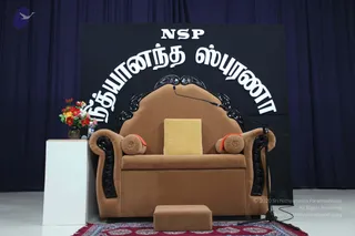 An empty brown upholstered throne-style chair with ornate black carved wooden arms and back stands on a stage before a dark blue curtain and a black backdrop. A yellow cushion rests on the seat, with orange-trimmed round bolsters at the sides and a footstool with a red patterned rug in front. A microphone on a stand curves toward the seat, and a white pedestal to the left holds a vase of red and yellow flowers and a water bottle. The backdrop displays the white Tamil word for a person Spurana above the letters NSP. No people are present.