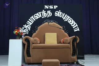 An empty brown velvet throne-style armchair with carved black wooden trim and orange-trimmed bolster cushions sits on a red patterned rug in front of a dark stage backdrop. A gold square cushion rests on the seat, a microphone on a stand is angled toward the chair, and a vase of red and yellow flowers stands on a white pedestal to the left. Above, white Tamil lettering reads NSP and the stage banner. No people are visible.