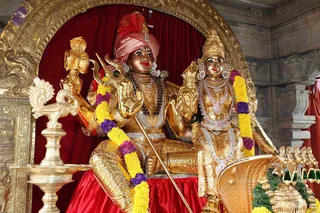 Golden murti of a seated deity pair under a gilded arch with red drapery. The male figure wears a pink and red turban, a rudraksha mala and gold necklaces, holds a trident and a staff, and is garlanded with yellow and purple flowers. The female figure wears a jeweled gold crown, a multi-strand necklace and ornate earrings and is also garlanded with yellow and purple flowers. A gold oil lamp stands at left and a golden vahana-style base is at lower right. No people are visible; this is a statue, so the people count is zero and the subject is not an in-person candidate.