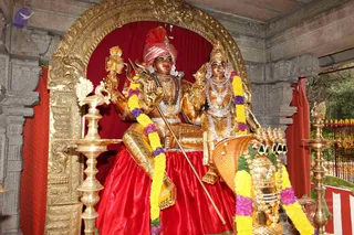 A photograph of a gilded deity murti pair seated together on a raised throne within an ornate golden arch, against a red cloth backdrop. The figure on the left wears a red and pink turban and holds a trident and a staff, with a rudraksha-style mala and gold necklaces; the figure on the right wears a gold crown, layered pearl and gold necklaces and earrings. Both are adorned with yellow and purple flower garlands. A brass oil lamp stands at the left and another at the right, and a gold vahana-like animal figure with a raised hood appears at the lower right. Stone pillars with carved motifs frame the scene.