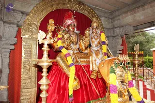 A photograph of a gilded murti of a seated deity couple within a golden arched niche draped with red fabric. The male figure wears a red and pink turban, a rudraksha-style garland and gold ornaments, holding a trident and a staff, and sits on a red cloth-covered seat. The female figure beside him wears a gold crown, layered white and silver necklaces and gold jewellery. Yellow and purple flower garlands hang over both figures. In front are a tall brass oil lamp and a golden kalasha-like vessel, with a golden cobra-hooded ornament at right. The setting is a stone temple structure with carved pillars and a green tree in the background. This is a deity murti, not a living person.