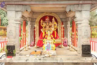A stone temple shrine with carved pillars and a gilded arched frame houses two golden deity murtis seated side by side on a raised platform, one a male figure with a red turban and the other a female figure with a tall crown, both draped in red garments. Yellow and white marigold garlands hang from their shoulders. Before them is a shivalinga-style stone form decorated with flower garlands, surrounded by brass oil lamps, a kalasha, small bowls and offering vessels. Red curtains frame the shrine and red-and-white railings enclose the area with green foliage behind.