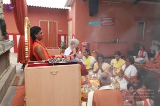 A photograph of a pada puja ceremony inside a red-walled temple hall. A man with long swept-back dark hair, wearing an orange robe and a rudraksha mala, sits on a golden-framed wooden throne at left, his hands resting in his lap. A woman in white with short grey hair and a red border, palms pressed together, kneels before him. Beside her, women in white and yellow shawls sit with folded hands, and a bald man in white and orange kneels at the lower foreground near brass plates of flowers and offerings. Incense smoke drifts through the room, and a red curtain hangs behind the throne. A sign on the back wall reads about annadanam.