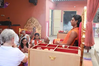 A seated man in an orange saffron garment with a swept-back dark hairstyle, sandalwood tilak on his forehead and red-and-white beaded malas around his neck, sits upright on a gold-and-red throne with hands folded, eyes closed. Before him stands a wooden offering box with compartments holding red apples and other fruit, and smoke rises from an inner doorway behind. Around him, devotees sit in an orange-walled hall: a woman in a pink sari with folded hands, a woman in an orange-and-white outfit, a bald man in white and a bald man in orange, one with a microphone raised near his mouth. A grey-haired woman in a white top sits in the foreground with palms together. An empty gold throne stands in the background beside a tripod-mounted camera.