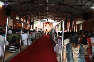 A photograph taken from the back of a covered hall with a red carpet runway leading toward a decorated altar at the far end. Rows of seated devotees fill both sides of the aisle, many in white or light clothing, with a woman in the foreground wearing a blue and cream patterned shawl over a floral kurta. Hanging cylindrical banners with red and blue patterns line the ceiling, and a red and white shrine with a deity murti is framed by pillars at the end of the hall. Speakers are mounted at the upper corners. The scene suggests a satsang or pada puja gathering, though no a person candidate is clearly visible.
