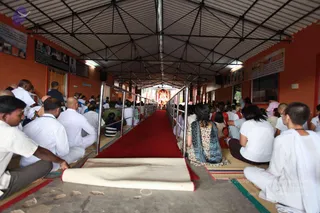 A long covered hall with orange walls and a corrugated roof, filled with seated devotees on mats facing a central red carpet aisle leading toward a distant shrine lit with red and gold decorations. Men on the left wear white shirts and white dhotis, some with shaved heads; a woman in a blue and floral dress and dupatta sits on the right, and women in white and dark clothing sit nearby. Metal railings line the aisle. Framed photographs hang on the orange walls, and a cream runner rolls across the foreground. Several dozen people are visible, many seated in rows facing the shrine.