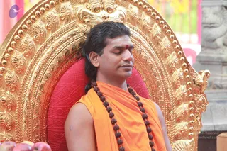 A man with swept-back long dark hair sits with eyes closed on an ornate gold throne with a red cushion. He wears a sleeveless saffron-orange wrap, a red kumkum dot on his forehead, and a long rudraksha mala of dark beads down his chest. Gold carved scrollwork and a stone pillar are behind him, with fruit visible at the lower left. The scene suggests a seated teaching or meditation moment. Swamiji (probable): the swept-back long hair, saffron wrap and rudraksha mala on a gold throne are the qualifying cues, so this is the seated Swamiji.