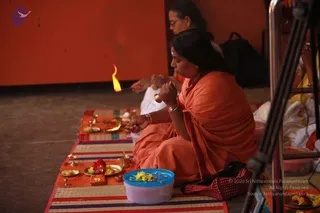 A woman in an orange sari sits cross-legged on a striped mat in a pada puja setting, holding her hands together near a small brass lamp with a lit flame. Her hair is pulled back and she wears gold bangles and a wristwatch. In front of her are brass plates with yellow and red flowers, a small brass cup, sweets and a blue container of marigold petals. Another woman in orange sits behind her, and an orange wall forms the backdrop. A red handbag rests on the mat at right.