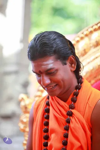 A smiling man with long dark hair swept back, wearing an orange sleeveless top draped with an orange shawl and a long rudraksha mala that hangs to his waist. He has a red bindi and white tilak on his forehead and looks down. Behind him is an ornate golden throne with round gold finials and a red cushion.