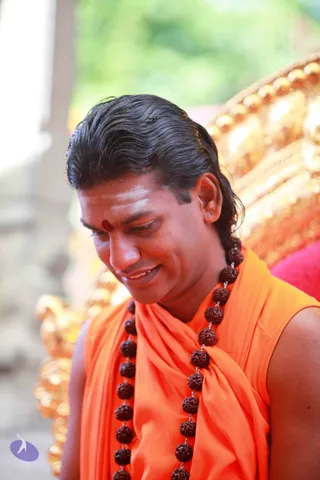 A smiling man with long dark hair swept back, wearing an orange sleeveless saffron wrap draped over the shoulders and a long strand of rudraksha beads. A red kumkum dot and white vibhuti marks adorn his forehead. He looks downward, seated in front of an ornate golden chair with a red cushion. A blurred green background. Swamiji (probable) appears in person, with cues of long swept-back hair, saffron clothing and rudraksha mala.