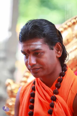 Close photo of a man with swept-back long dark hair, white ash lines on the forehead and a red kumkum dot, wearing an orange saffron shawl over a sleeveless orange garment and a long rudraksha mala with dark brown beads. He looks downward with a faint smile. Behind him, blurred golden ornamental decor and green foliage. Swamiji (probable): long swept-back hair, saffron clothing and rudraksha mala, shown in person.