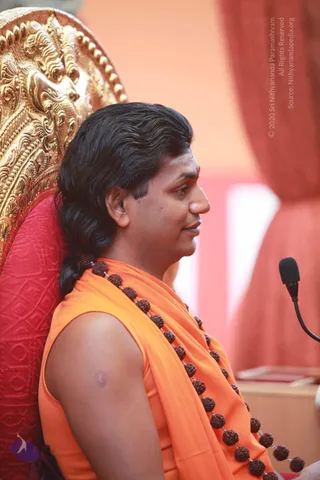 A man in profile sits on a red cushioned seat with an ornate gold-carved throne backrest behind him. He wears a sleeveless saffron-orange garment draped over the shoulder and a long strand of dark rudraksha beads across his chest. His long dark hair is swept back and tied, and he has a red tilak on his forehead. A black microphone on a flexible stand is at the right edge. The background is soft coral-pink drapery. Probable Swamiji is seated in a ceremonial chair, with his saffron clothing, rudraksha mala and long swept-back hair as visible cues.