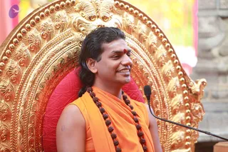 A man seated on an ornate gilded throne with a red cushion, speaking into a microphone on a stand at right. He wears a sleeveless saffron-orange garment draped over his shoulders, a long rudraksha mala with multiple strands of brown beads hanging to his waist, and a red bindi on his forehead. His long dark hair is swept back and he smiles warmly. Swamiji (probable): the long swept-back hair, saffron clothing, rudraksha malas and seated throne are visible cues of the Swamiji candidate. The setting appears to be an outdoor satsang with a pink backdrop behind the throne.