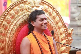 A man with long dark hair swept back sits in front of an ornate gold throne with a red cushion, smiling toward the side. He wears an orange sleeveless top with an orange shawl draped over his shoulders, a long necklace of rudraksha beads, and a red bindi on his forehead. A black microphone stand is in the lower right foreground. Behind him, a stone wall and soft pink and yellow background blur. The appearance is consistent with the Swamiji (probable) cues of long swept-back hair, orange clothing, and rudraksha mala, seated on a throne at a satsang talk.