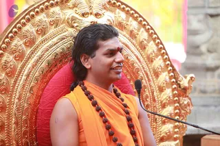 A man seated on an ornate gold throne with a red cushion, wearing an orange sleeveless garment and a long dark rudraksha mala, with a red tilak on his forehead and hair swept back into a low tail. A black microphone on a stand is in front of him. Swamiji (probable): long swept-back hair, saffron clothing and rudraksha mala on a throne. The ornate gold backrest and a blurred pink and green backdrop are visible behind him.