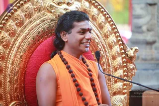 A smiling man with long dark hair swept back sits on an ornate gold throne with a red cushion. He wears an orange sleeveless garment with a shawl draped over one shoulder and a long strand of brown rudraksha beads, and a red bindi on his forehead. A microphone on a stand is in front of him. a person appears as the seated speaker, based on the long swept hair, saffron clothing, rudraksha mala and throne. The background shows a stone pillar and blurred greenery.