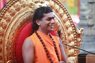 A man seated on an ornate gold throne with a red cushion, smiling. He wears an orange sleeveless garment with a draped orange shawl, a long rudraksha mala around his neck, and a red bindi on his forehead. His dark hair is long and swept back. A black microphone on a stand is in front of him, and a blurred green and red background with a stone pillar is behind. Swamiji (probable): long swept-back hair, orange clothing, rudraksha mala and ornate gold throne suggest the Nithyananda Paramashivam, seated as a speaker during a satsang talk.