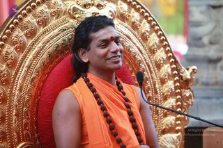 A smiling man with long dark hair swept back sits on an ornate gold throne with a red cushion. He wears an orange sleeveless garment with a shawl draped over one shoulder, a long rudraksha mala with dark beads hanging to his chest, and a red bindi on his forehead. A microphone on a stand is in front of him. Golden repousse carving fills the background of the throne, and a stone wall is visible at right.
