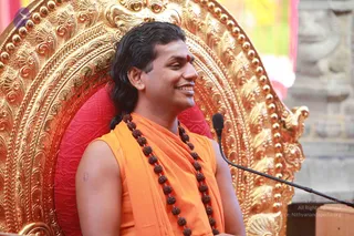 A smiling man with long dark hair swept back sits on an ornate gilded throne with a red cushion. He wears a sleeveless saffron-orange garment over his shoulders and a long necklace of brown rudraksha beads, and has a red bindi on his forehead. A microphone on a stand is in front of him, and a wooden lectern edge is visible at lower right.