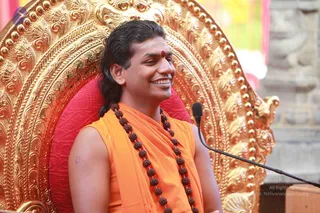 A smiling man with long dark hair sits on a red cushion before an ornate gold throne with repoussé floral and scroll carvings. He wears a sleeveless saffron-orange garment over his shoulders, a long necklace of dark rudraksha beads, and a red bindi on his forehead. A microphone on a stand is at his right, and a pink fabric draping and stone pillar appear in the background. Appearance-based label: a person, seated and speaking as a teacher at a satsang.