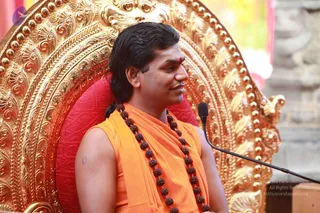 A man seated on an ornate gilded throne with a red cushioned back, speaking into a black microphone on a stand at right. He wears an orange sleeveless top with a sash-like orange wrap, a long rudraksha mala with large brown beads, and a red tilak on his forehead; his dark hair is swept back. The gilt carved backdrop and red cushion dominate the frame.