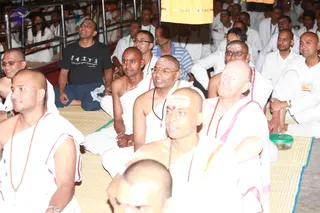 A seated crowd of devotees at an evening satsang, sitting on woven mats with a large yellow cloth hanging at the top edge. Several shaved-headed men wear white dhoti-style garments with pink or magenta borders and rudraksha malas; some have white and red tilak marks on their foreheads. In the centre, a man in white with rudraksha beads and a white-and-red forehead mark smiles while listening. Behind them, a man in a black t-shirt stands, and others in white and blue sit among the audience. The scene conveys an attentive public talk with devotees listening closely.