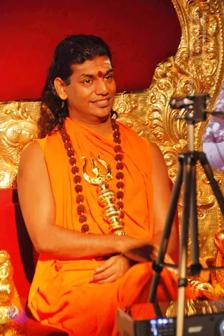 A man seated on an ornate gold throne against a red backdrop, wearing an orange saffron wrap and a long rudraksha mala with a gold trident pendant, with a red bindi on his forehead and long swept-back dark hair. His hands are clasped in his lap. A tripod-mounted camera and microphone stand are in the foreground at right. Swamiji (probable): the man is a seated spiritual teacher, identified by long hair, saffron clothing and rudraksha malas as the Swamiji, giving a satsang talk.