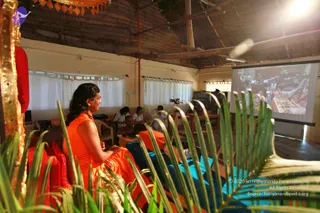 A photograph of a seated woman in an orange sleeveless top and gold-trimmed orange garment, long dark hair, sitting on a red cushioned seat beside a gilded frame at the left edge. Palm fronds in the foreground. Behind her, several people sit at tables with laptops in a thatched-roof hall with white curtains. On the right, a projector screen shows a crowded gathering with an altar-like display.
