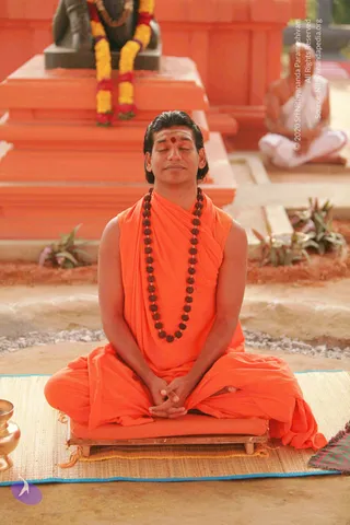 A man in orange robes sits cross-legged with eyes closed and hands folded in his lap on a wooden seat over a woven mat, in meditation. He wears a long rudraksha mala, a red bindi and white tilak, and his hair is swept back. Behind him, an orange stepped pedestal holds a black stone deity murti garlanded with yellow and red flowers. A second seated figure in white sits in the background at right, and a brass vessel rests at the lower left.