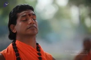Close-up photo of a man with long, swept-back dark hair and a red kumkum dot on his forehead, eyes closed in meditation. He wears an orange saffron shawl draped over bare shoulders and a thick necklace of dark rudraksha beads. The background is a soft blur of green foliage, with another figure in orange robes blurred at the lower right.