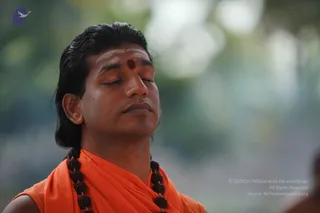 Close-up photograph of a man with swept-back dark hair, eyes closed and face lifted in meditation, wearing a red bindi on his forehead, an orange saffron shawl wrapped over his shoulders, and a dark rudraksha mala around his neck. Blurred green foliage fills the background. A small figure is partly visible at the lower right edge. The man appears to be a person, given his long swept-back hair, saffron wrap and rudraksha mala, though his face is not used for identification.