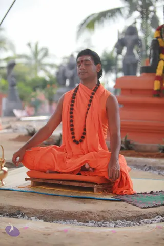 A man sits cross-legged in meditation on a low wooden platform with an orange cushion, set on a woven mat with a blue edge outdoors. He wears orange robes with a sleeveless top and an orange wrap over one shoulder, a dark rudraksha mala around his neck, and a red-and-white tilak on his forehead; his eyes are closed and his hands rest on his knees. Behind him stands a large orange pedestal and a garland of yellow and red flowers, with grey stone statues and palm trees in the background. A brass kalasha sits at the left edge and gravel borders the mat. Appearance-based cues (long swept-back hair, saffron robes, rudraksha mala) suggest a probable Swamiji (probable) seated in meditation, consistent with the page's talk.