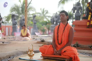 A photograph of a man seated cross-legged in meditation on a low wooden platform covered with a cushion, placed on a woven mat. He wears orange saffron robes, a sleeveless orange garment draped over the shoulders, and a long rudraksha mala, with a red tilak on his forehead and hair swept back. Eyes are closed and hands rest folded in his lap. A brass kalasha-style vessel with a handle sits on the mat to his left. Behind him stands a large orange plinth with a garland of yellow and red flowers, and dark stone statues and sculptures are arranged in a garden. A second person in a yellow shawl and pale clothing sits further back, partly visible. The setting is an outdoor ashram courtyard with palm trees and sandy ground.