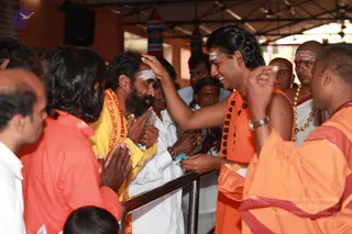 A photograph of a darshan moment at a crowded hall with a metal railing. A man in a long-haired, swept-back style wearing orange robes and a rudraksha mala, with a white forehead mark, smiles while placing his hand on the forehead of a bearded devotee with a white tilak. The devotee wears a yellow and red shawl over white clothes and holds a card and folded hands. Other devotees in white and orange stand around, and a shaved-headed priest in orange with a gold-bead necklace is at right. Swamiji (probable): the orange-robed man with long swept-back hair, rudraksha mala and ceremonial dress is the candidate, giving blessing to a devotee.
