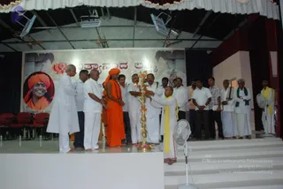 A photograph of an indoor stage event with a large brass multi-tier oil lamp (aarti lamp) being lit at centre, adorned with flowers. Standing around it are several men in white dhotis and white shirts, a man in a yellow shawl kneeling to tend the lamp, and a priest in white with a yellow shawl and a tilak. Beside them stands a figure in saffron-orange robes with a saffron head wrap and a rudraksha mala, the cues consistent with the a person presentation. Behind them a large banner shows a portrait of a smiling figure in an orange turban and robes, with Kannada script above, and a lotus motif. Red chairs sit at the left, and a standing fan and stage steps are visible at the right.