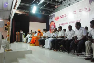 A stage event in a hall with a white stage platform, a wooden podium on the left, and a brass oil lamp with orange marigold garlands beside it. Seated on the stage in a row are a person in orange robes with an orange head covering on an orange chair, and several men in white shirts and dark trousers or white dhotis. A man in a white shirt stands at the podium speaking. A large banner behind them shows a portrait of a smiling person in an orange headcloth beside a logo and the Kannada text. Audience members in white shirts sit in chairs at the right edge. A white pedestal fan stands at the lower left.