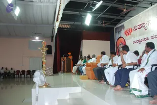 Wide photograph of an indoor stage hall with a white draped ceiling and ceiling fans. A standing speaker in an orange-saffron robe with an orange head wrap stands at a wooden podium with a microphone, facing the audience. Seated on the right side of the stage are rows of men in white dhotis and white shirts, several in dark blue shirts, and one in a green shawl. A tall brass oil lamp with orange flower garlands stands at the left of the stage. A large banner behind the stage shows a portrait of a smiling man in an orange head wrap and the a person logo, with Kannada script text. A white floor fan sits in the foreground. Several people sit in red chairs at the left edge.
