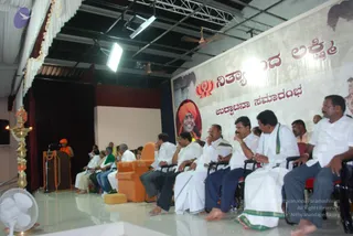 Photograph of a stage event at a hall with white draped curtains and a brown wooden podium with a microphone. A figure in orange robes stands at the podium, speaking. Seated in a row of chairs in front are men in white dhotis and white shirts, several with bare feet, some holding their hands to their faces while listening. A large orange-highlighted armchair sits center stage. A brass oil lamp with a lit flame stands at the left edge. A backdrop banner shows a portrait of a man in an orange turban with the Kannada-script caption reading Nithyananda Lakshmi and the word for inauguration. An orange-robed figure at the podium may be a sannyasi giving a talk.
