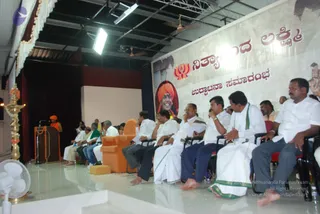 A photograph of a stage event in a large hall with white ceiling fans and stage lights overhead. On the left, a speaker in an orange robe stands at a wooden podium draped with orange and white garlands, beside a brass oil lamp. A large banner behind the stage shows a portrait labelled "Nithyananda Lakshmi" with a saffron-turbaned figure, and on the right a lit deity murti. Seated in rows of chairs in the foreground are several men in white dhotis and white shirts, some with green-bordered dhotis, several wearing wristwatches and sitting in an audience of roughly a dozen visible people. A large orange armchair sits center stage. The floor is polished white.
