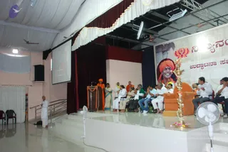 Photograph of a stage event in a hall with white ceiling drapes, maroon curtains and a projection screen. A banner on the right shows a portrait labelled with Kannada text and a lotus emblem, depicting a smiling figure in an orange-red turban. A standing man in white clothes and white dhoti stands at the left edge near a railing. At a wooden podium draped with marigold garlands, a person in orange robes stands beside a woman in a teal sari. Seated on the stage are about a dozen people in white and pastel clothes, several with hands folded in greeting. An empty orange armchair sits beside a large brass oil lamp (aarti lamp) in the foreground. Two standing fans are visible at the lower corners.
