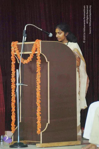 A woman in a cream saree with a gold border stands at a brown wooden podium on a stage, reading or speaking into a silver microphone on a flexible stand. Orange marigold garlands drape the podium. Dark maroon velvet curtains hang behind her, and a white-clad figure is partly visible at the right edge.