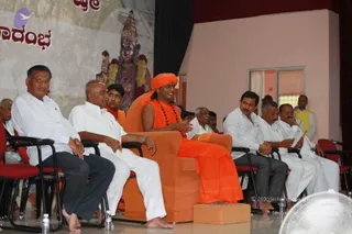 A photograph of a seated public talk on a stage. In the centre, a person in saffron-orange robes and an orange head wrap sits in an orange armchair with a rudraksha mala around the neck, holding papers and speaking, with an orange footstool before them. Flanking them on both sides are men in white shirts and trousers and dark trousers seated in red-cushioned chairs, some holding papers. Behind the stage is a painted backdrop with a red-and-white deity image garlanded with flowers. Water bottles stand on the floor and a fan is in the lower right corner. Several other people sit in the background.