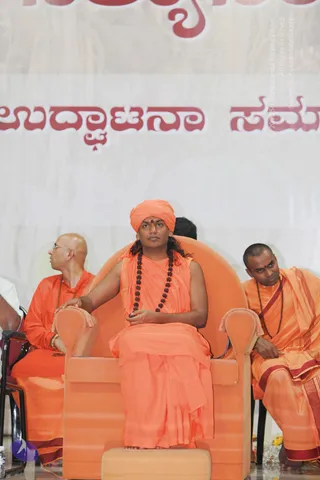 A seated man in an orange saffron turban with a red tilak on his forehead sits in an orange upholstered armchair on a stage. He wears a sleeveless orange robe over a long-hanging orange garment and a dark rudraksha mala draped around his neck, with long dark hair falling past his shoulders. His right hand rests on the armrest. To his left, a bald man in orange robes and wire-rimmed glasses looks off to the side, and to his right, a man with a shaved head and orange clothes with maroon borders sits in a chair wearing a rudraksha mala and a red tilak. Behind the stage is a large white banner with red Kannada script. A bouquet of yellow flowers sits at the lower right edge. The seated central figure matches the Swamiji (probable) cues of long swept hair, a saffron turban, saffron robes and rudraksha beads, and is shown in person on stage.