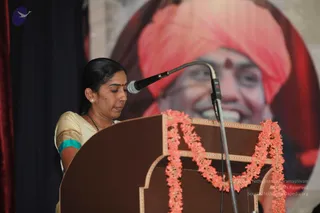 A woman stands at a brown wooden lectern with a microphone on a flexible stand, her head lowered as she speaks. She wears a cream and teal sari with gold border, a small red bindi, a dark beaded necklace and pearl earrings, and her hair is pulled back. Orange-pink flower garlands drape the lectern. Behind her is a large printed portrait of a smiling man in a pink-red turban with a red bindi, shown only as a picture rather than a physical person. A dark curtain frames the left side.