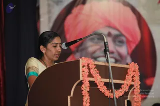 A woman in a cream and teal sari with a red bindi stands at a wooden lectern speaking into a microphone, with an orange-pink garland draped over the lectern. Behind her a large backdrop shows a smiling man in a pink-orange turban with a red forehead mark, a depiction of a portrait rather than a person present on stage. A dark curtain frames the left side and a small blue logo appears in the upper left corner.