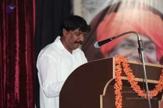 A man in a white shirt stands at a dark wooden podium with his head bowed, speaking into a silver-stemmed microphone. Orange marigold garlands hang along the podium edge. Behind him, a large portrait shows a smiling figure in a saffron-orange turban and crimson robes, set against a deep red velvet curtain and dark backdrop. The man is a speaker or presenter at a public talk; the portrait is a printed depiction on the backdrop.