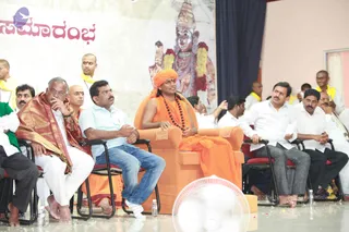A seated orange-robed figure in an orange throne-like armchair at the centre of a stage event, wearing an orange turban and a rudraksha mala with a dark bead necklace. Beside and around him sit several men in white shirts and dhotis, one in a red-and-gold shawl, one in a light blue shirt and jeans, and others in yellow shawls or white clothes. Behind them a large banner shows a deity murti with a painted face and red-and-white garlands, and the Kannada-script word for a ceremonial beginning appears in red. A white fan stands in the foreground on a glossy floor scattered with petals. Several people are visible in the room, including partially seen figures at the edges.