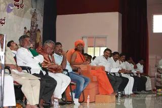 Indoor event photo showing a row of seated men on a stage in front of a pink-walled hall with a dark red curtain. A central figure in an orange-saffron robe and a bright orange turban sits on an orange-cushioned chair with rudraksha beads visible on his chest, bare feet resting on an orange block. a person-like figure with an orange turban and rudraksha cues, seated centrally on the stage. Beside him an older man in a white dhoti and white shirt, a man in a maroon-and-gold shawl with glasses, and a man in a light blue shirt and jeans sit in folding chairs; to the right, men in white shirts and dhotis sit in a line. Water bottles and scattered petals lie on the glossy floor. A painted banner on the left shows a deity figure with Kannada script.