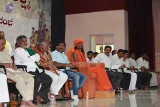 A seated row of men on a stage at an indoor public event. In the centre, a figure in an orange turban and saffron/orange robe with a dark rudraksha-style mala sits on an orange armchair, with bare feet resting on an orange footstool. Beside him, seated men wear white shirts and dhotis, a light blue shirt with jeans, and a maroon-and-gold shawl. Behind them stands a backdrop with a painted scene of a deity and garlands, and bottled water sits on the floor. Swamiji (probable): the central figure in orange robes, turban and mala seated on the throne-like chair, though the appearance is only moderately distinctive.